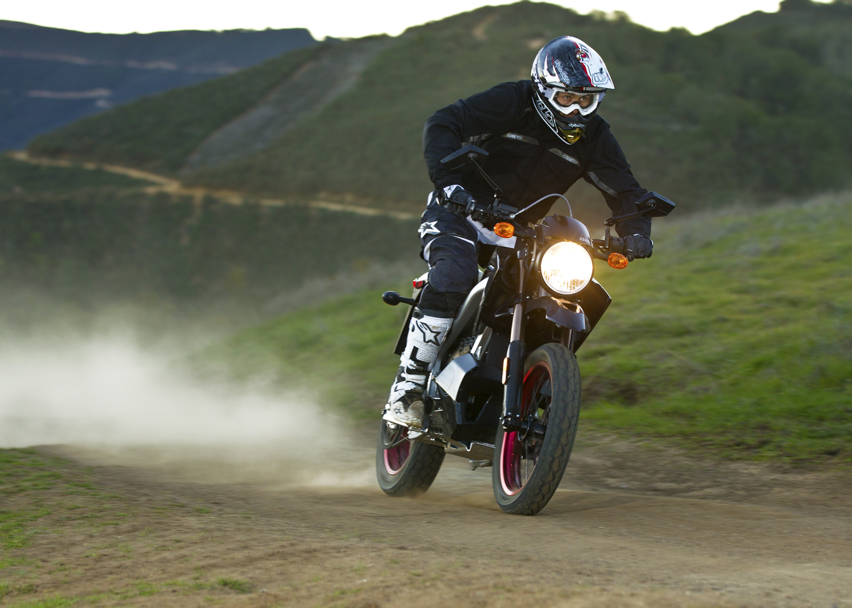 Man riding an electric motorcycle with motocross style helmet, goggles and shin guards on a dirt track on a mountain trail.